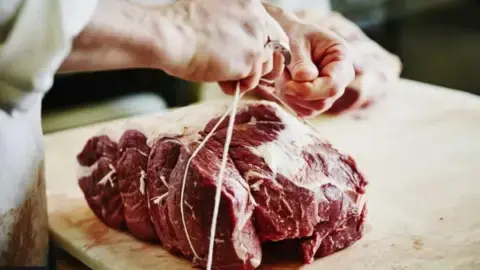 Getty Images A large chunk of red meat sitting on a butchers block. A person wearing a white coat is tying white string around it.