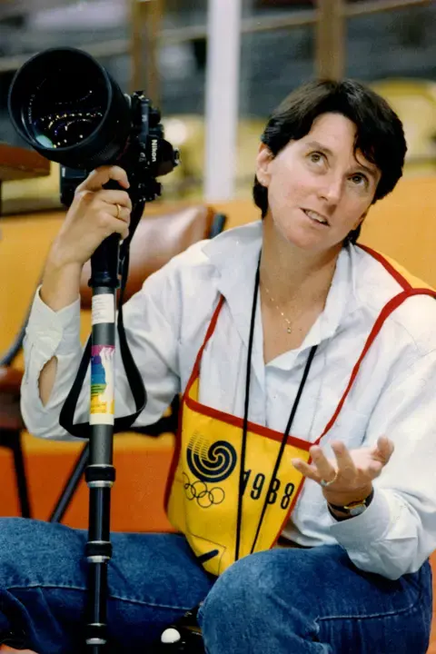 Eileen Langsley Eileen sitting on a stool with her camera. She is wearing an Olympic media vest.