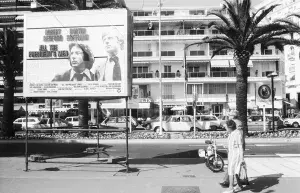 An eldery couple walking down a sidewalk in Cannes, France and looking at a billboard advertisement for "All the President's Men" starring Robert Redford and Dustin Hoffman, a parking lot, hotel, and palm trees visible in the background (Photo by Guy Marineau/WWD/Penske Media via Getty Images)