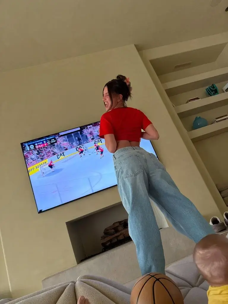 Hailey Bieber posing in front of a TV wearing light-wash jeans with a red slogan tee, which she tied to give a cropped silhouette.