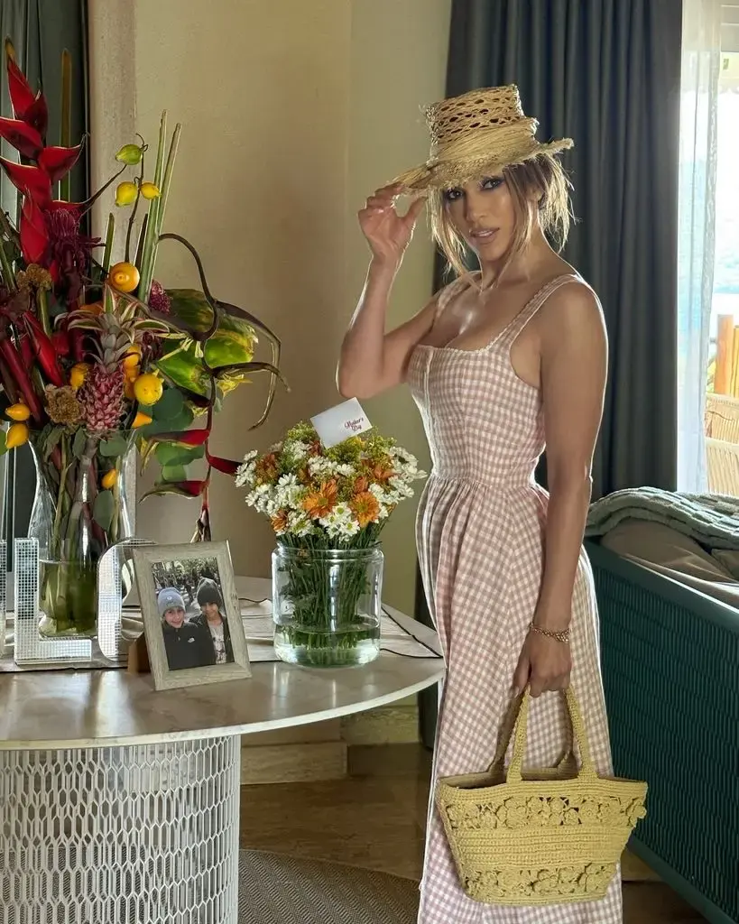 In this warmly lit indoor photograph, Jennifer Lopez stands gracefully beside a round table adorned with vibrant floral arrangements and a framed photograph of two children. She wears a soft pink and white gingham milkmaid-style dress that hugs her figure and flares gently at the bottom. Her hair is styled in relaxed waves and partially tucked under a wide-brimmed raffia hat. On her arm, she carries a straw-like raffia tote bag with intricate floral crochet detailing. The ambiance is breezy and summery, with natural light streaming in from a window in the background, highlighting the seaside setting beyond the room. Her expression is serene, radiating a casual elegance.