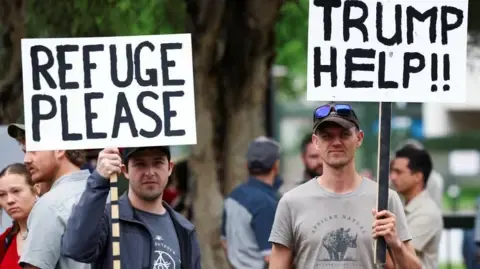 Reuters Two demonstrators hold placards saying "REFUGE PLEASE" and "TRUMP HELP!!" outside the American Embassy in Pretoria, South Africa in February